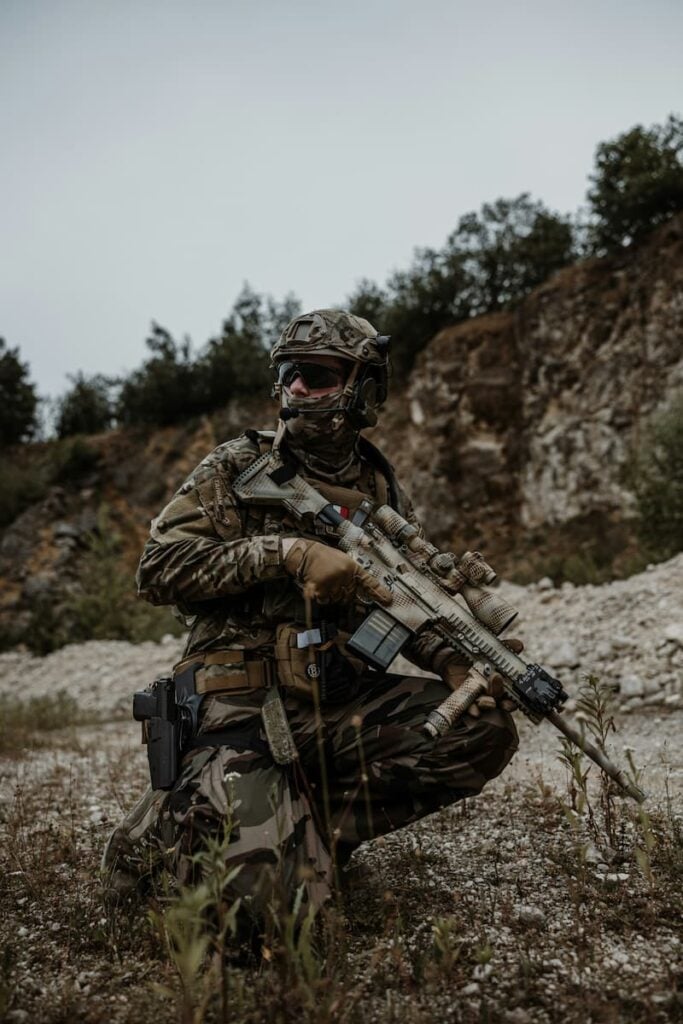 Soldier operating in rocky terrain wearing tactical equipment and carrying a rifle during a military exercise.