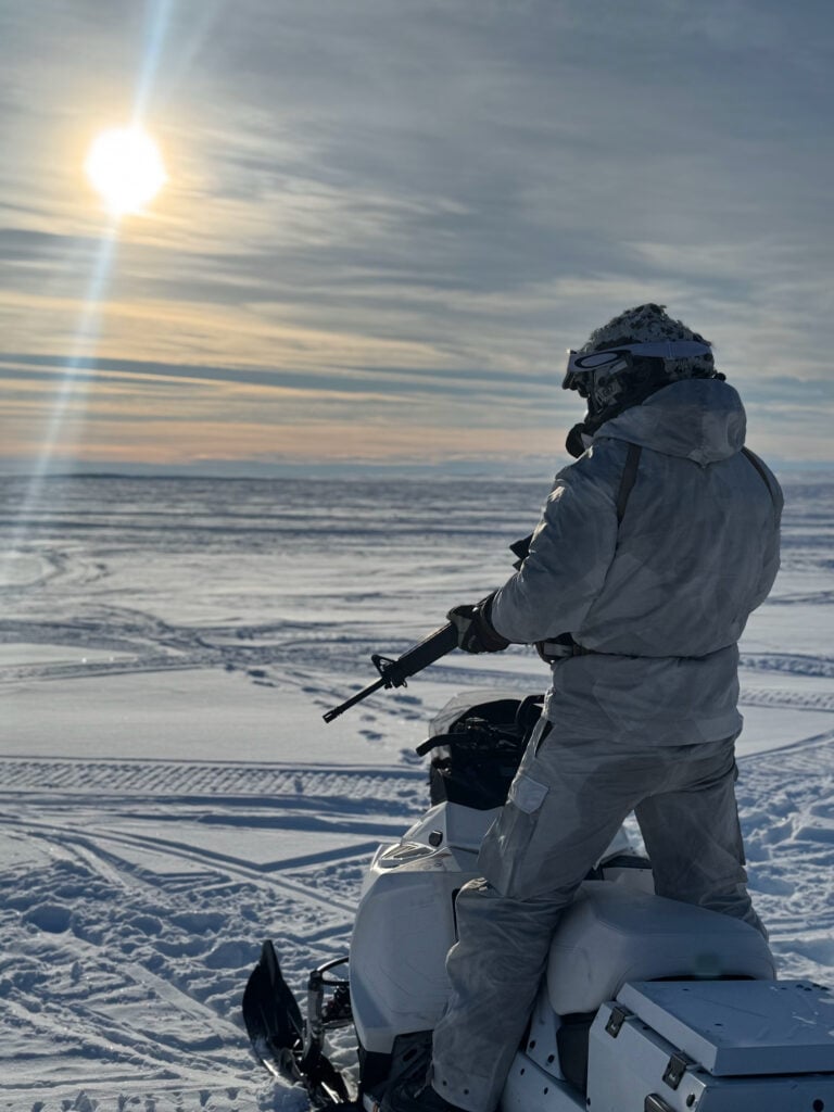 Soldier in white cold-weather clothing carrying a rifle during operations in a snowy Arctic environment.